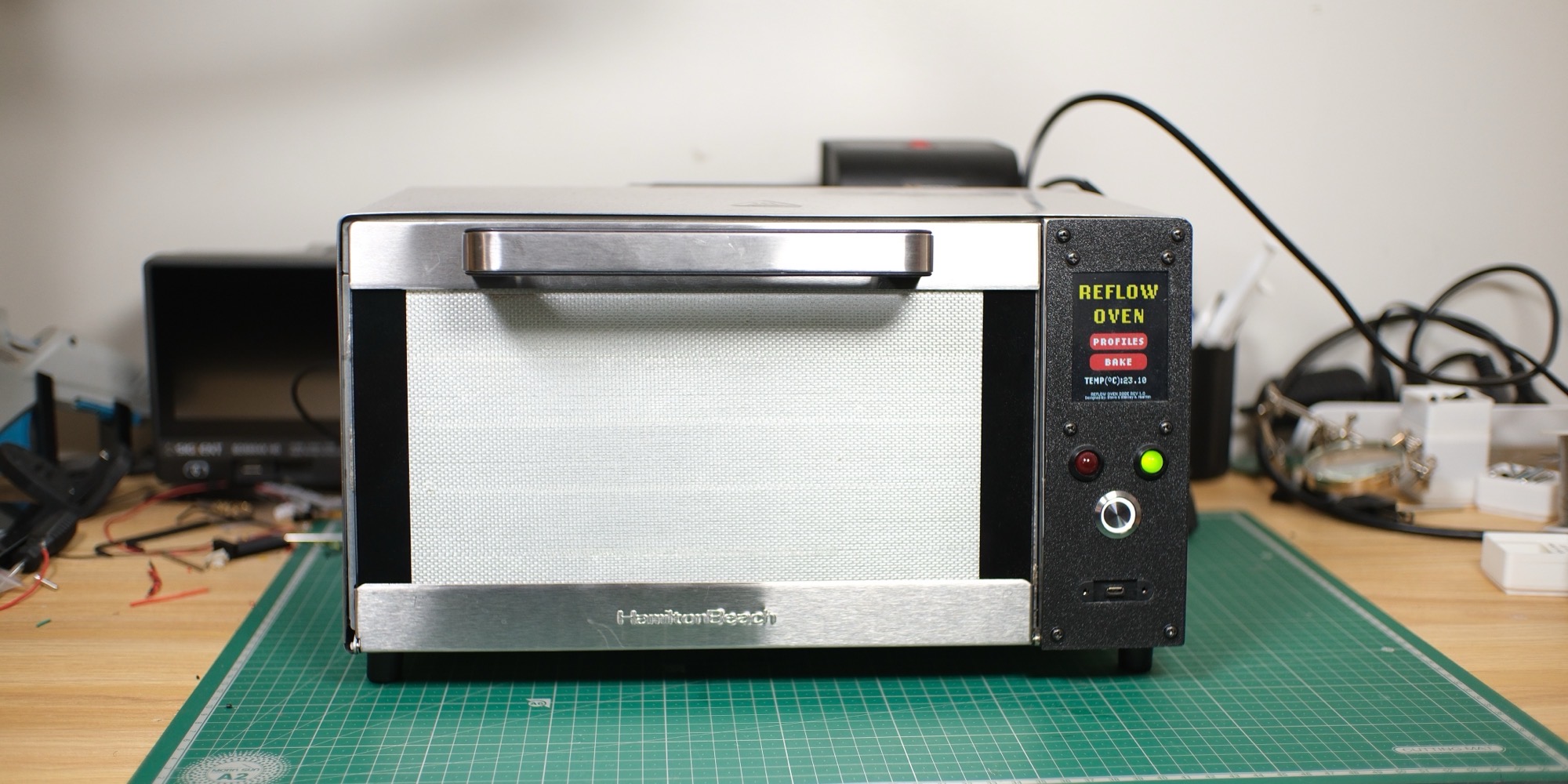 The reflow oven on a cutting mat: a Hamilton Beach toaster oven with a black 3D-printed face plate holding the touchscreen, two LEDs and a switch
